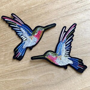 Puede incluir: Dos parches bordados de colibríes. Los parches presentan costuras detalladas en tonos de azul, rosa, verde y blanco, con contornos negros. Los colibríes están representados en pleno vuelo, con las alas extendidas.
