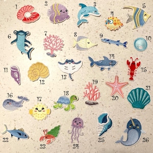 Può includere: Una collezione di toppe ricamate raffiguranti varie creature marine e vita marina. Include pesci, un delfino, uno squalo, un'aragosta, una stella marina, una tartaruga, una balena, una medusa e coralli.
