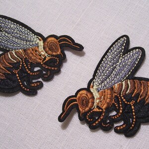 May include: Two embroidered bee patches. The bees have brown and black striped bodies, tan heads, and detailed wings with grey and white accents. The patches are outlined in black.
