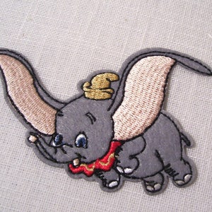 以下が含まれることがあります： グレーの象がピンクの大きな耳、赤と金の襟、金の帽子をかぶっている刺繍のアイロン接着パッチ。象は鼻を伸ばして飛んでいます。