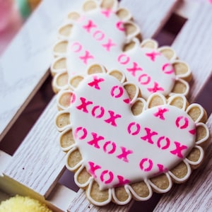 May include: Two heart-shaped sugar cookies decorated with white icing and pink "XO" patterns. The cookies are on a white wooden surface.