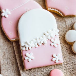 May include: Pink and white sugar cookies decorated with white icing flowers. The cookies are shaped like a dress, a heart, and a flower.