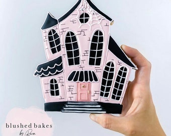 May include: A pink and black gingerbread house cookie decorated with white icing and black details. The house has a spiderweb on the front and a black door with a gold knob.