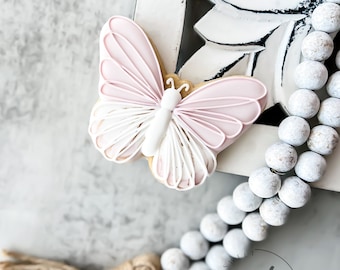 May include: A pink and white butterfly cookie decorated with royal icing. The cookie is sitting on a white wooden surface with a string of white beads and a brown tassel.