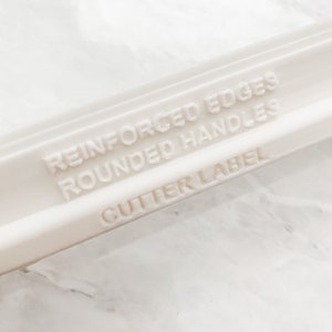 May include: White plastic rolling pin with the text "Reinforced Edges Rounded Handles Cutter Label" embossed on the side.