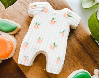 May include: A white baby onesie cookie decorated with orange and green icing. The onesie has a striped pattern and is decorated with small orange circles. The cookie is on a wooden surface.