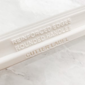 May include: Close-up of a white plastic object with the text "REINFORCED EDGES, ROUNDED HANDLES, CUTTER LABEL" embossed on its surface. The object appears to be a tool or part of a tool, possibly for crafting or baking.