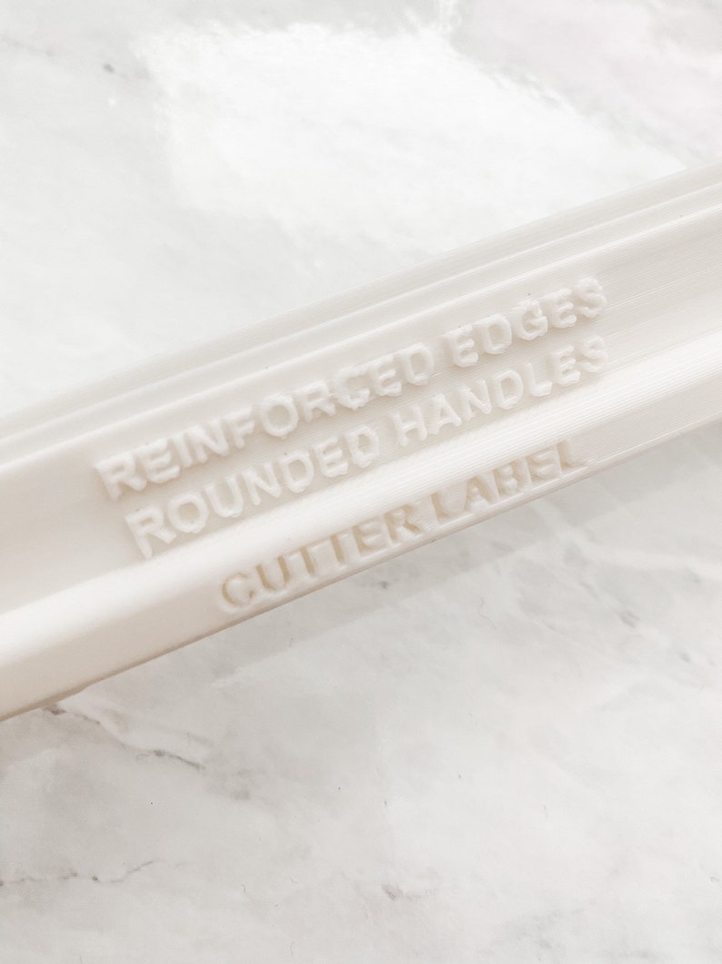 May include: Close-up of a white plastic object with the text "REINFORCED EDGES, ROUNDED HANDLES, CUTTER LABEL" embossed on its surface. The object appears to be a tool or part of a tool, possibly for crafting or baking.