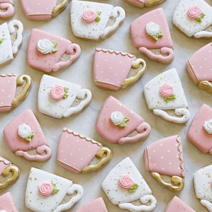 May include: A collection of pink and white teacup-shaped biscuits decorated with gold accents and white flowers. The biscuits are arranged on a white surface.
