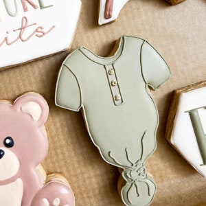 May include: Cookies decorated with pastel-colored icing. A baby onesie cookie with gold button details is the focal point. Other cookies include a teddy bear, a sign with the word "Future" and a rocking horse.