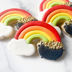 May include: Four rainbow-colored sugar cookies decorated with a black pot of gold at the end of the rainbow. The cookies are on a white marble surface.