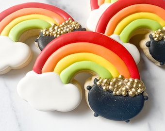 May include: Four rainbow-colored sugar cookies decorated with a black pot of gold at the end of the rainbow. The cookies are on a white marble surface.