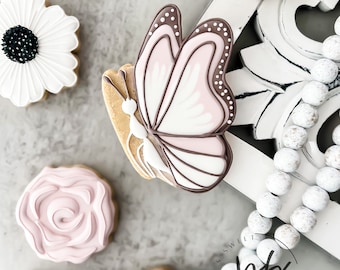 May include: A collection of decorated sugar cookies featuring a pink rose, a white daisy, a pink butterfly, and a white flower with a black center. The cookies are arranged on a white surface with a white beaded garland and brown tassels.