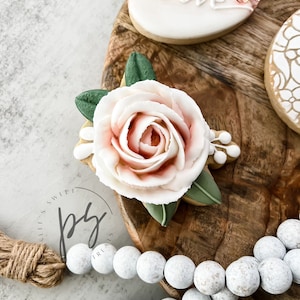 May include: Close-up of decorated sugar cookies. One cookie features a large, light pink and white flower with green leaves. Another cookie has the word "love" written on it. The cookies are on a wooden surface.
