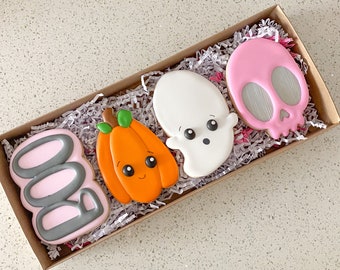 May include: A set of four Halloween-themed cookies in a brown box with shredded paper. The cookies are decorated with pink icing and gray icing. The cookies are shaped like a ghost, a pumpkin, a skull, and the word "BOO".