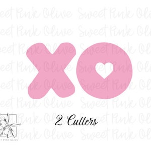 May include: Two pink cookie cutters, one shaped like an 'X' and the other shaped like a circle with a heart inside. The text '2 Cutters' is printed below the cutters.