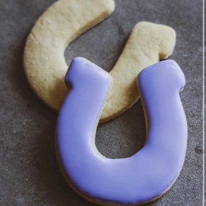 May include: Two sugar cookies shaped like horseshoes. One horseshoe is plain and the other is decorated with purple icing.