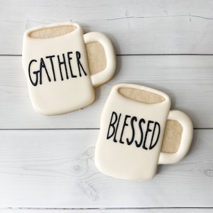 May include: Two mug-shaped cookies with white icing and beige filling. One cookie has the word "GATHER" in black, the other says "BLESSED". The cookies are on a white wooden surface.