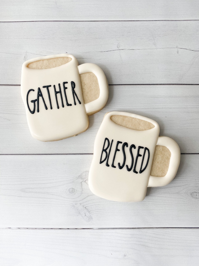 May include: Two mug-shaped cookies with white icing and beige filling. One cookie has the word "GATHER" in black, the other says "BLESSED". The cookies are on a white wooden surface.