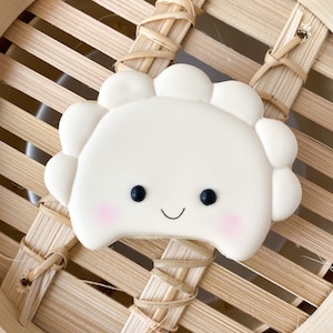 May include: A white cookie shaped like a dumpling with a smiling face and black eyes. The cookie is sitting in a bamboo steamer.