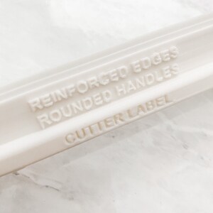 Puede incluir: Rodillo de pl&aacute;stico blanco con el texto "Reinforced Edges Rounded Handles Cutter Label" grabado en el lateral.