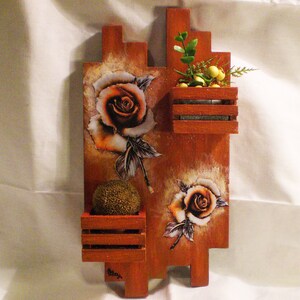 Peut inclure: Une étagère murale en bois avec trois compartiments, chacun présentant un motif floral différent. Le compartiment supérieur a une petite plante en pot avec des fleurs jaunes, le compartiment du milieu a une plante succulente et le compartiment inférieur a une grande rose.