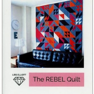 May include: A colorful quilt with geometric shapes in shades of red, blue, black, and white hangs on a wall in a modern living room. The quilt is titled "The REBEL Quilt" and is by Libs Elliott.