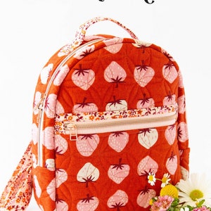 May include: A small orange quilted backpack with a strawberry print and a zipper closure. The backpack has a top handle and adjustable straps. The text "THE VIOLET BACKPACK" is at the top of the image. The text "KNOT + THREAD design" is at the bottom of the image.