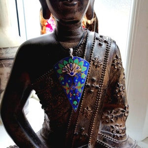 May include: A blue, green and gold patterned pendant with a silver chain hanging from the neck of a dark brown Buddha statue.