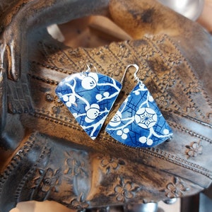 May include: A pair of blue and white ceramic earrings with a floral design. The earrings are shaped like a fan and have a silver hook.
