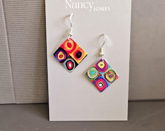 May include: A pair of colorful, abstract geometric earrings. Each earring is a square with four smaller squares, each containing a circle design in shades of red, yellow, blue, and green. The earrings are displayed on a gray card with the name "Nancy Jones".