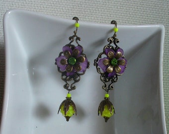 Könnte beinhalten: Ein Paar violette und grüne Blumenohrringe mit gelben Akzenten. Die Ohrringe sind aus Metall und haben einen Vintage-Stil.