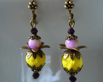 Könnte beinhalten: Ein Paar Hängeohrringe mit einer gelben und violetten Perle, einem bronzefarbenen Blumendesign und einem violetten Kristall am unteren Ende.