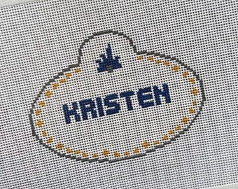 May include: A white needlepoint canvas featuring a stitched name tag design. The oval-shaped border is outlined in grey with yellow accents. Centered is the name KRISTEN in dark blue block letters, topped with a small blue castle icon.