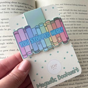 May include: A magnetic bookmark featuring a row of pastel-colored books with the text YOU STOPPED HERE! in blue. The bookmark is attached to a white backing card with light blue polka dots and the text JDP & Co. and Magnetic Bookmark.