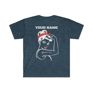 May include: A navy blue t-shirt with a white silhouette of a woman with red polka dot headband and the text "YOUR NAME" on the back. The woman is flexing her bicep and has a small American flag tattoo on her arm with the text "Sisters in Arms".