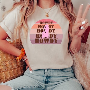 Peut inclure: Un t-shirt blanc avec un motif graphique brun et rose représentant un coucher de soleil et le texte "HOWDY" répété cinq fois.