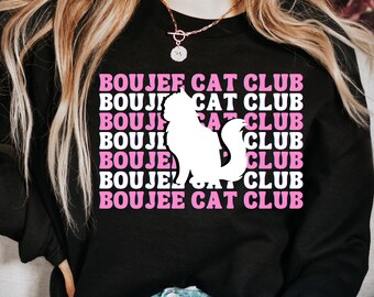 以下が含まれることがあります： ピンクの「BOUJEE CAT CLUB」の文字が繰り返し書かれた、猫の白いシルエットが描かれた黒いスウェットシャツです。
