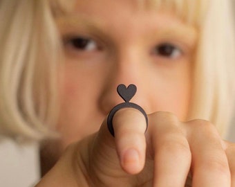 May include: A black ring with a heart-shaped cutout on top. The ring is worn on a finger.
