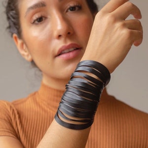 Puede incluir: Un brazalete de cuero negro con múltiples tiras finas de cuero envueltas alrededor de la muñeca.