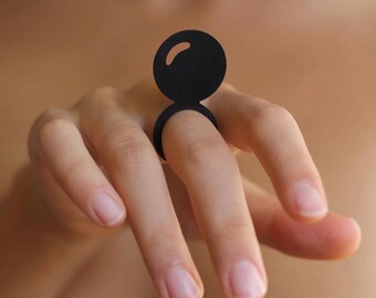 May include: A black ring with a large, round, and slightly raised top. The ring is worn on the index finger of a hand.