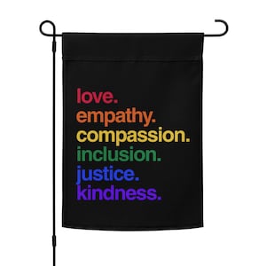 May include: Black garden flag with the words "love. empathy. compassion. inclusion. justice. kindness." in rainbow colors. The flag is attached to a black metal pole with a curved top.