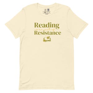 May include: Cream-colored t-shirt with the text "Reading is an act of Resistance" in a green-yellow font. The design includes an open book with floral accents. The shirt has a crew neck and short sleeves.