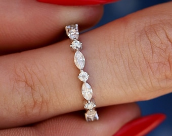 Puede incluir: Un anillo de banda en tono plateado con un patrón alterno de piedras transparentes de talla marquesa y redonda. Las piedras están engastadas en una fila continua a lo largo de la banda metálica, creando un diseño delicado y simétrico con acabado pulido.