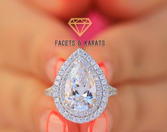 Puede incluir: Un anillo de plata con un diamante en forma de pera, rodeado por un doble halo de piedras más pequeñas. El anillo es sostenido por una persona con esmalte de uñas rosa. El logotipo de Facets & Karats está en la parte superior central.