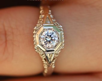 May include: A silver engagement ring with a round, clear diamond set in a hexagonal bezel. The ring has a delicate, intricate design with small diamonds set around the center stone.