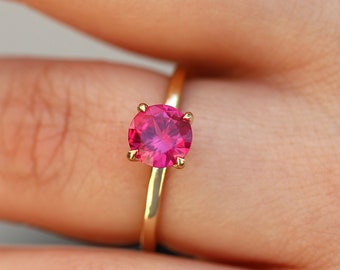 May include: A gold ring with a pink gemstone. The ring is on a finger.