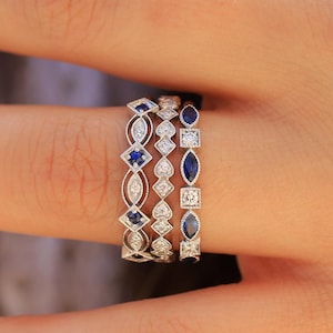 May include: Three delicate silver rings with alternating blue sapphires and clear diamonds. The rings are stacked on a finger.