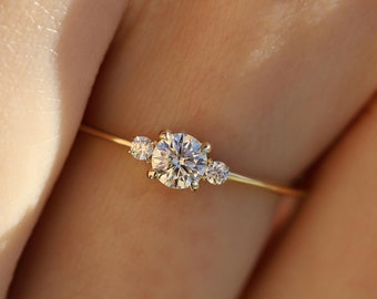 May include: A delicate gold ring featuring a central round diamond flanked by two smaller diamonds. The ring is set on a thin gold band, creating a minimalist and elegant design. The ring is shown on a hand, highlighting its size and sparkle.