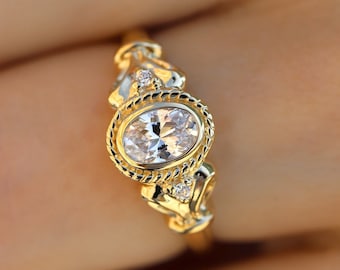 May include: A gold ring with an oval-shaped diamond set in a bezel setting. The ring has a detailed design with small diamonds on the sides.